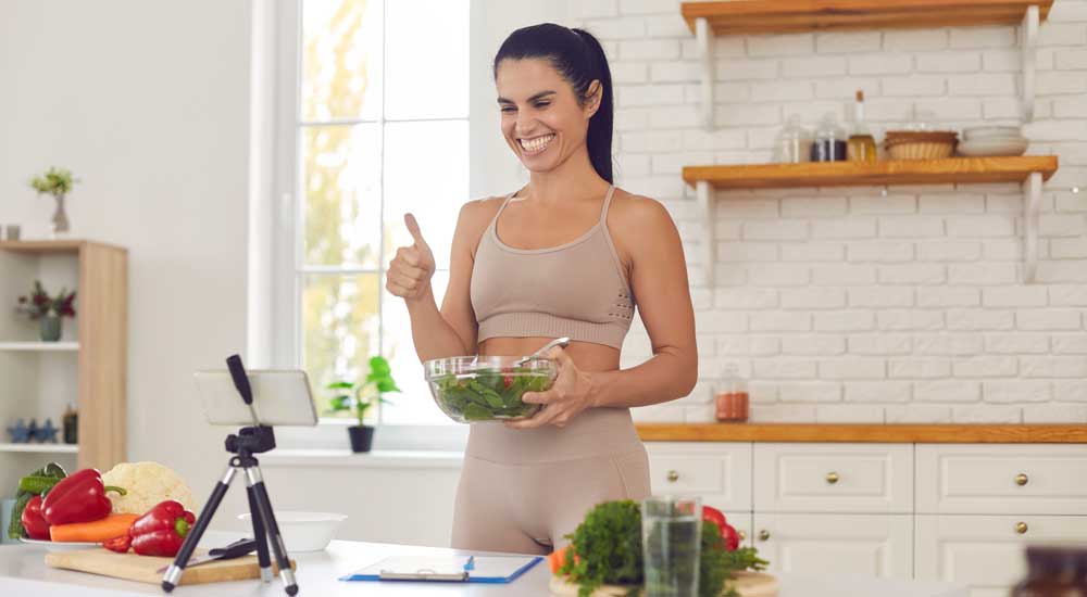 woman in kitchen holding salad bowl with phone recording