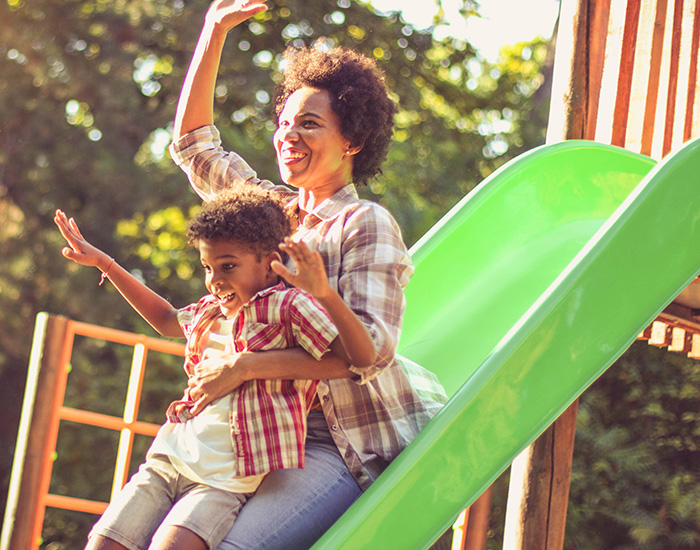 A mother and child smile as they play on a slide in a park.